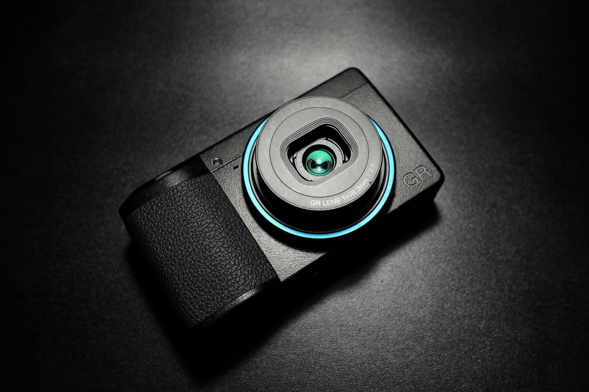 Ricoh GR III prémium kompakt fényképezőgép kék objektívgyűrűvel. Fekete Ricoh GR III kompakt fényképezőgép felülnézetből, kék gyűrűvel az objektív körül, sötét háttér előtt.