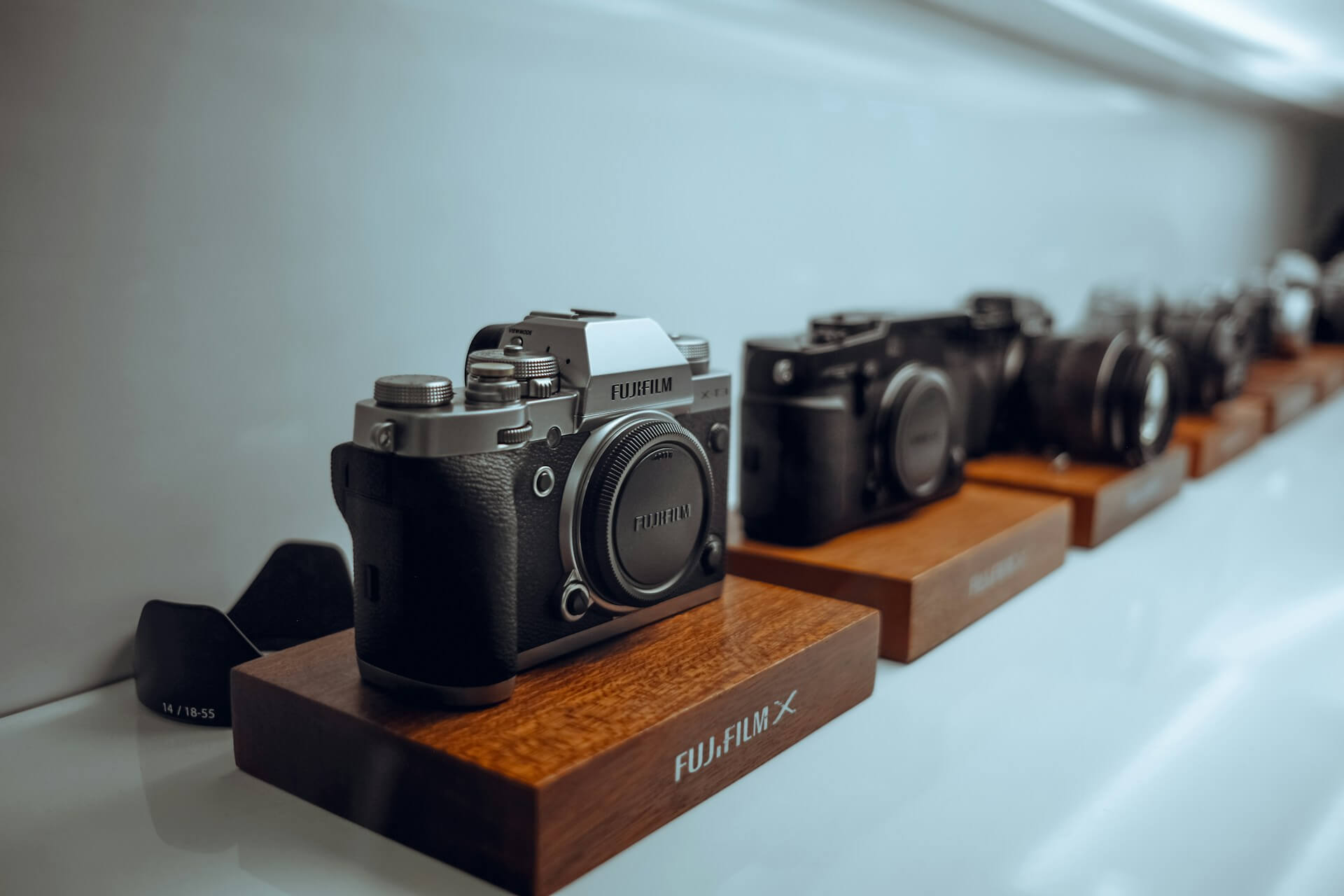 Fujifilm X-sorozatú fényképezőgépek bemutatóállványon. Faállványokon sorakozó Fujifilm X-sorozatú tükör nélküli fényképezőgépek, előtérben egy ezüst-fekete vázas filmmásoló retro dizájnnal.