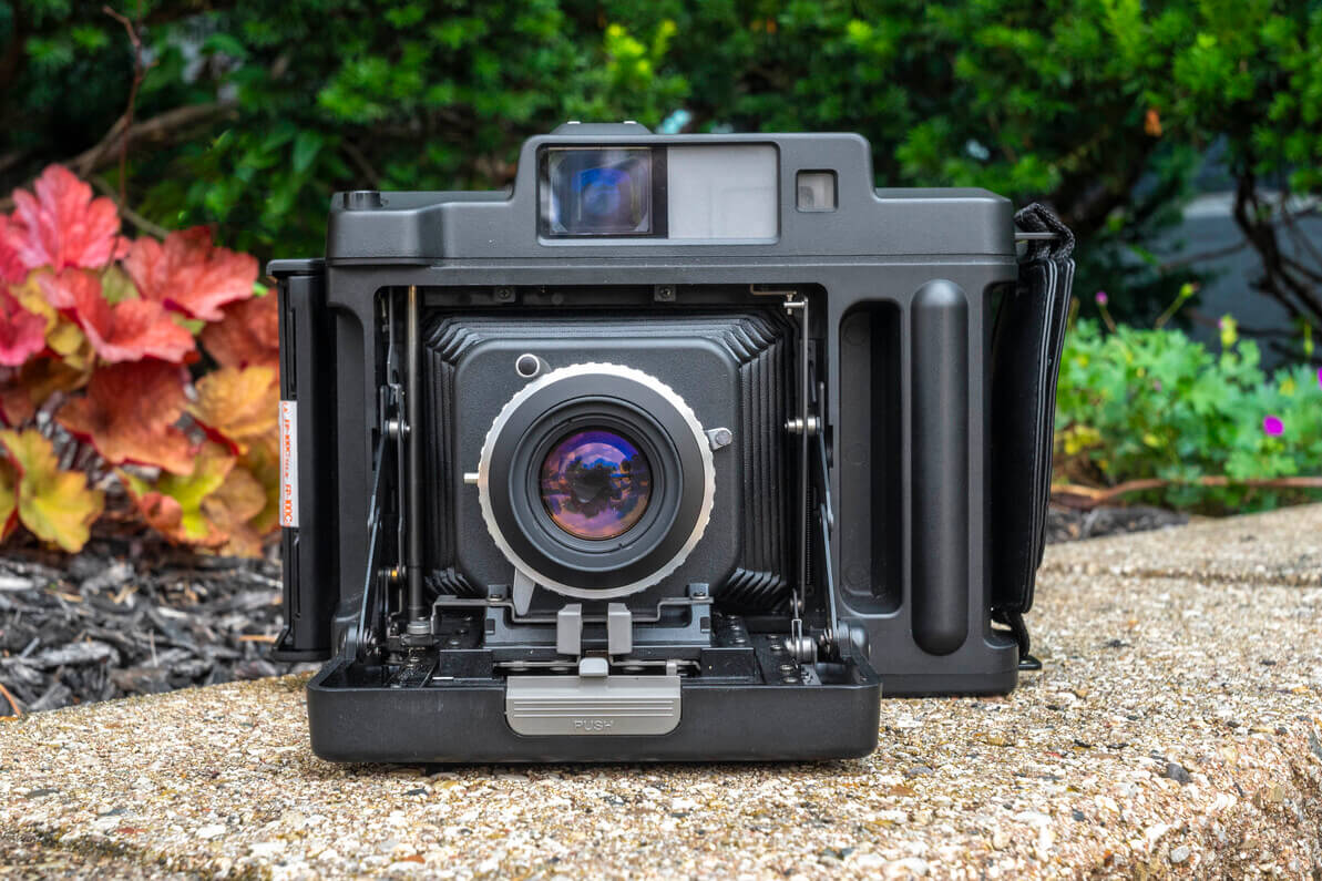 Szemből készült fotó egy kinyitott, fekete Fujifilm FP-1 Professional kihúzható harmonikás instant fényképezőgépről, kültéri háttérben.