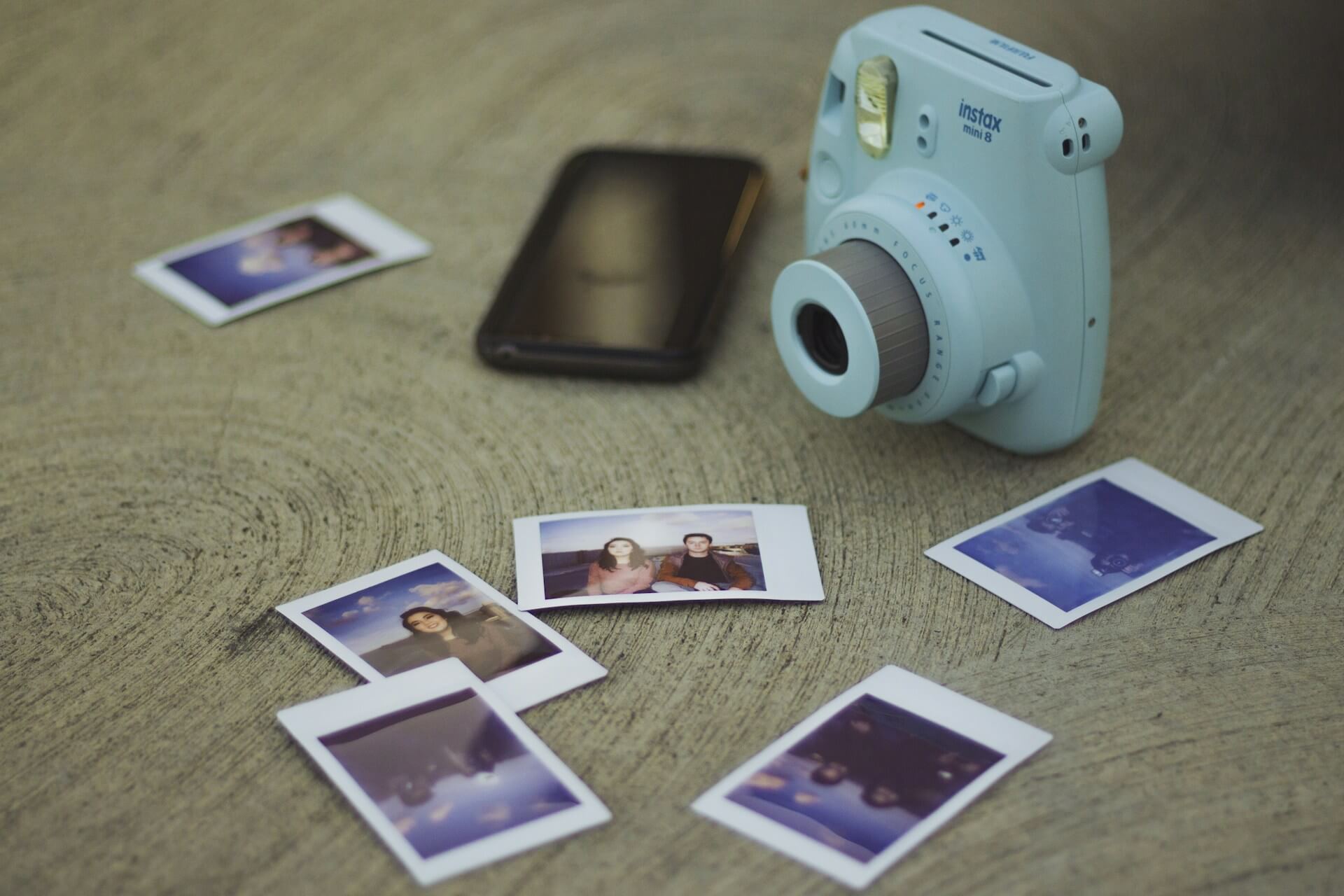 Világoskék Fujifilm Instax Mini 8 fényképezőgép és kis méretű instant fotók. Felülnézeti fotó egy asztalon lévő világoskék Fujifilm Instax Mini 8 instant fényképezőgépről, egy okostelefonról és szétszórt Instax fotókról.