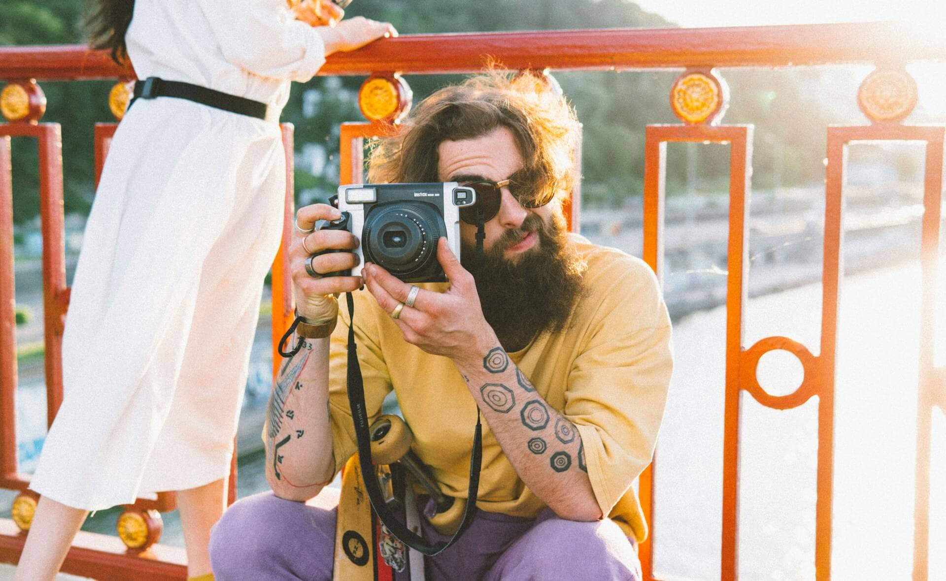 Tetovált, szakállas férfi napszemüvegben, amint egy Fujifilm Instax Wide fényképezőgéppel fotóz egy piros korlátú hídon, naplementében.