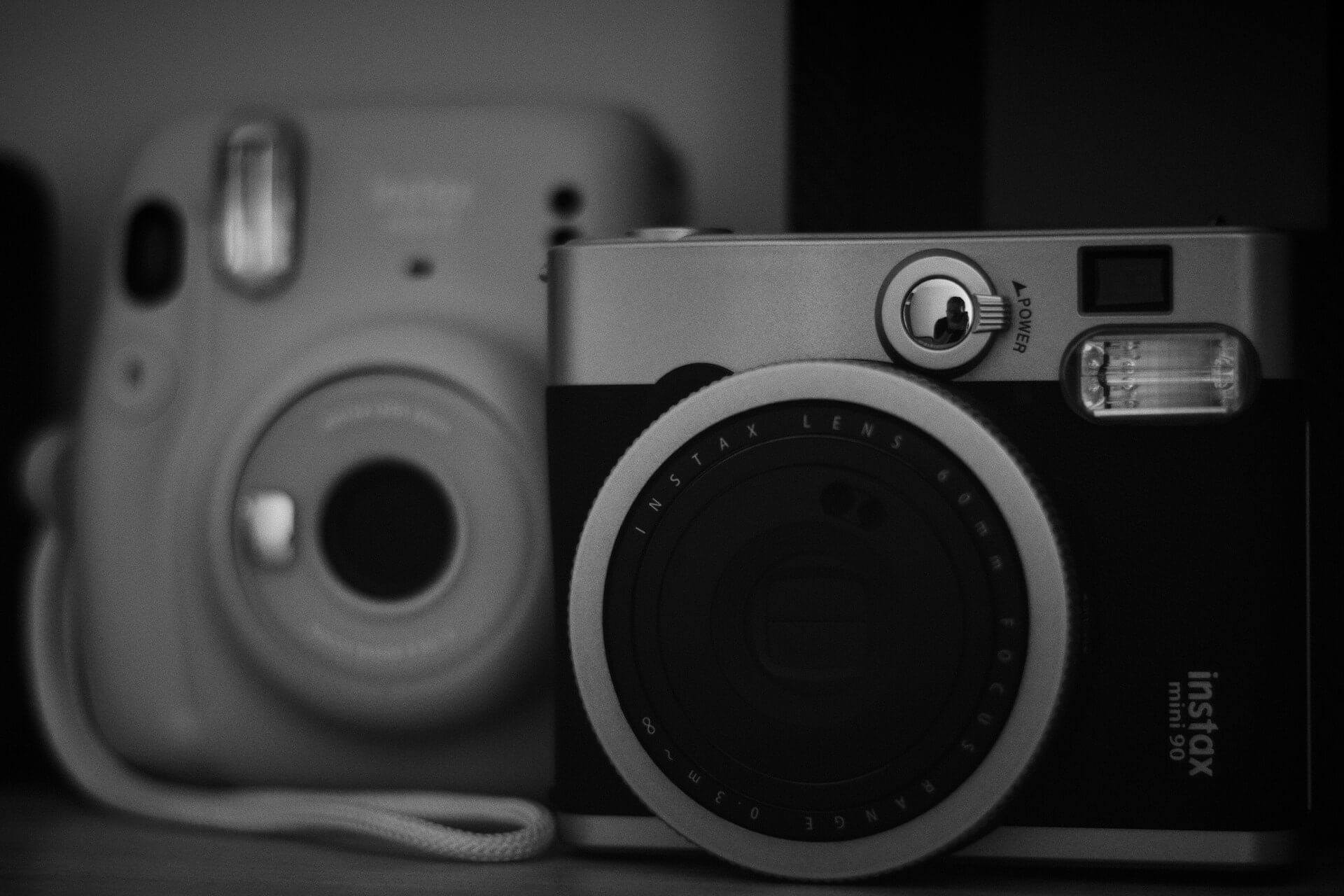 Retro Fujifilm Instax Mini 90 és Instax Mini 11 fekete-fehérben. Fekete-fehér fotó egy előtérben lévő retro stílusú Fujifilm Instax Mini 90 Neo Classic és egy háttérben homályosabban látszó Instax Mini 11 instant fényképezőgépről.