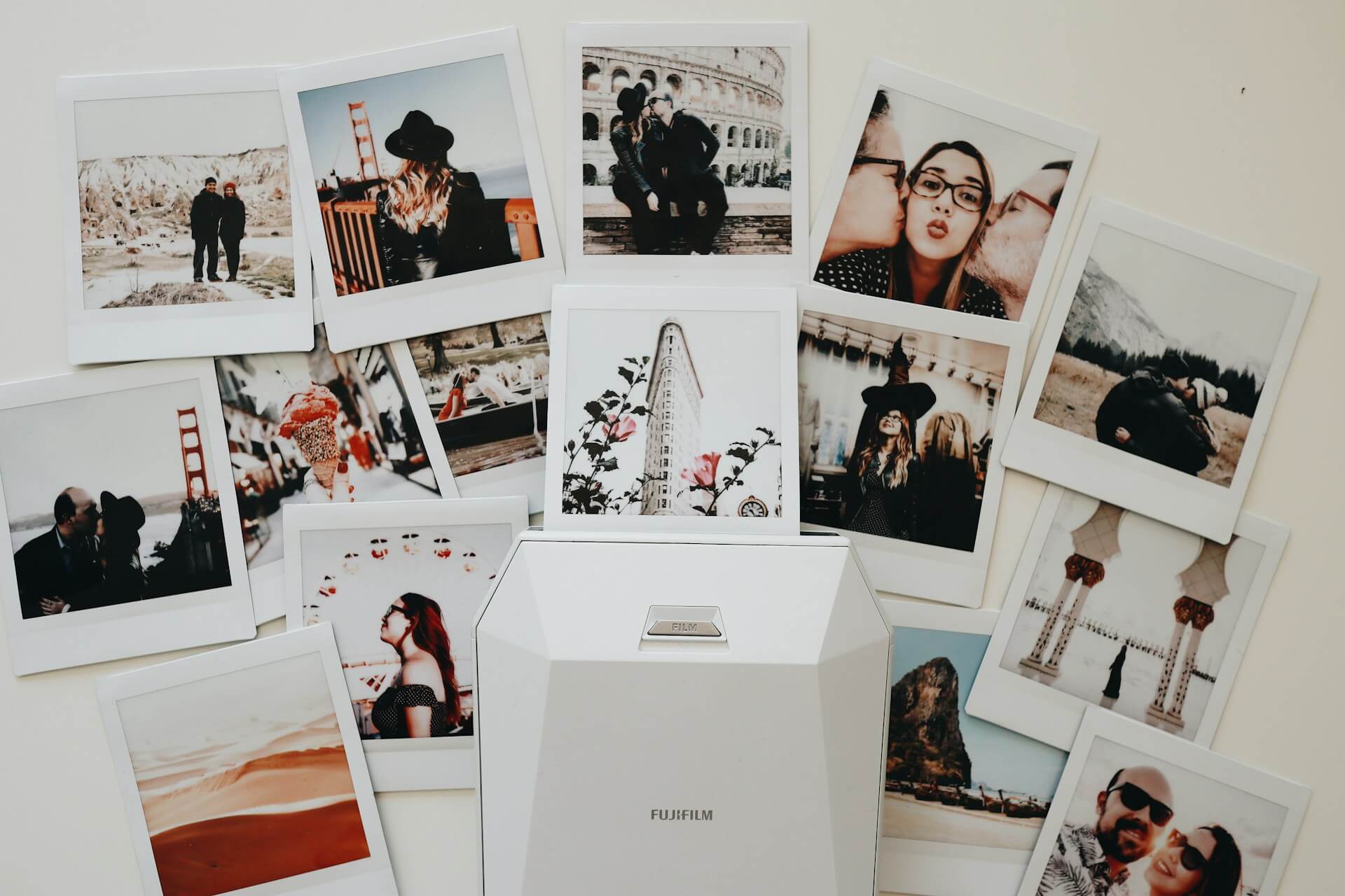 Fehér Instax fotónyomtató és kiterített instant képek. Fehér Fujifilm Instax SP-2/Share okostelefon-nyomtató, amelyből egy kép jön ki, körülötte sok, élményeket és portrékat ábrázoló instant fotóval.