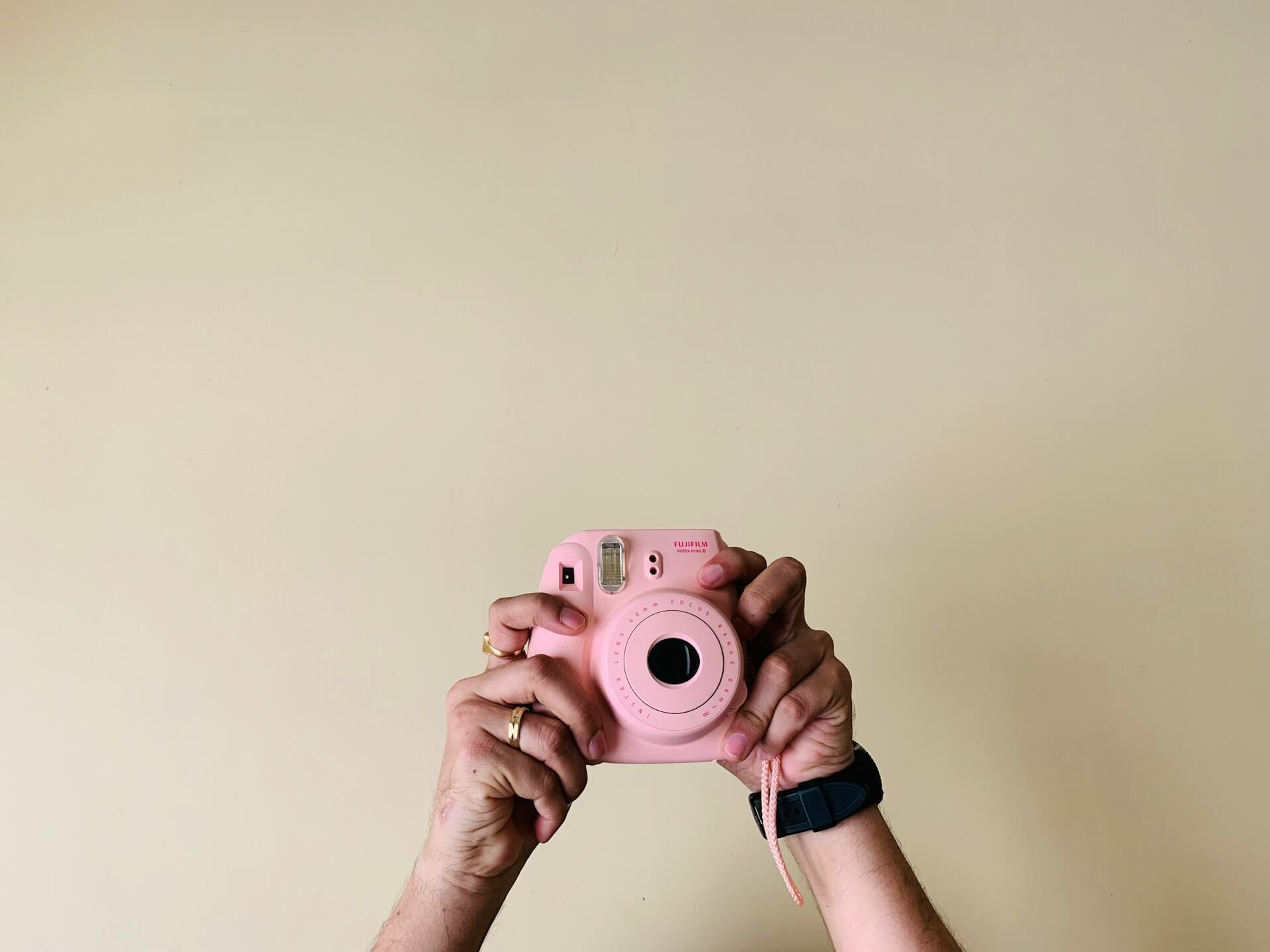 Rózsaszín Instax Mini 8 fényképezőgép kézben. Két kézzel magasba tartott, pasztellrózsaszín Fujifilm Instax Mini 8 fényképezőgép szemből, letisztult, bézs háttér előtt.