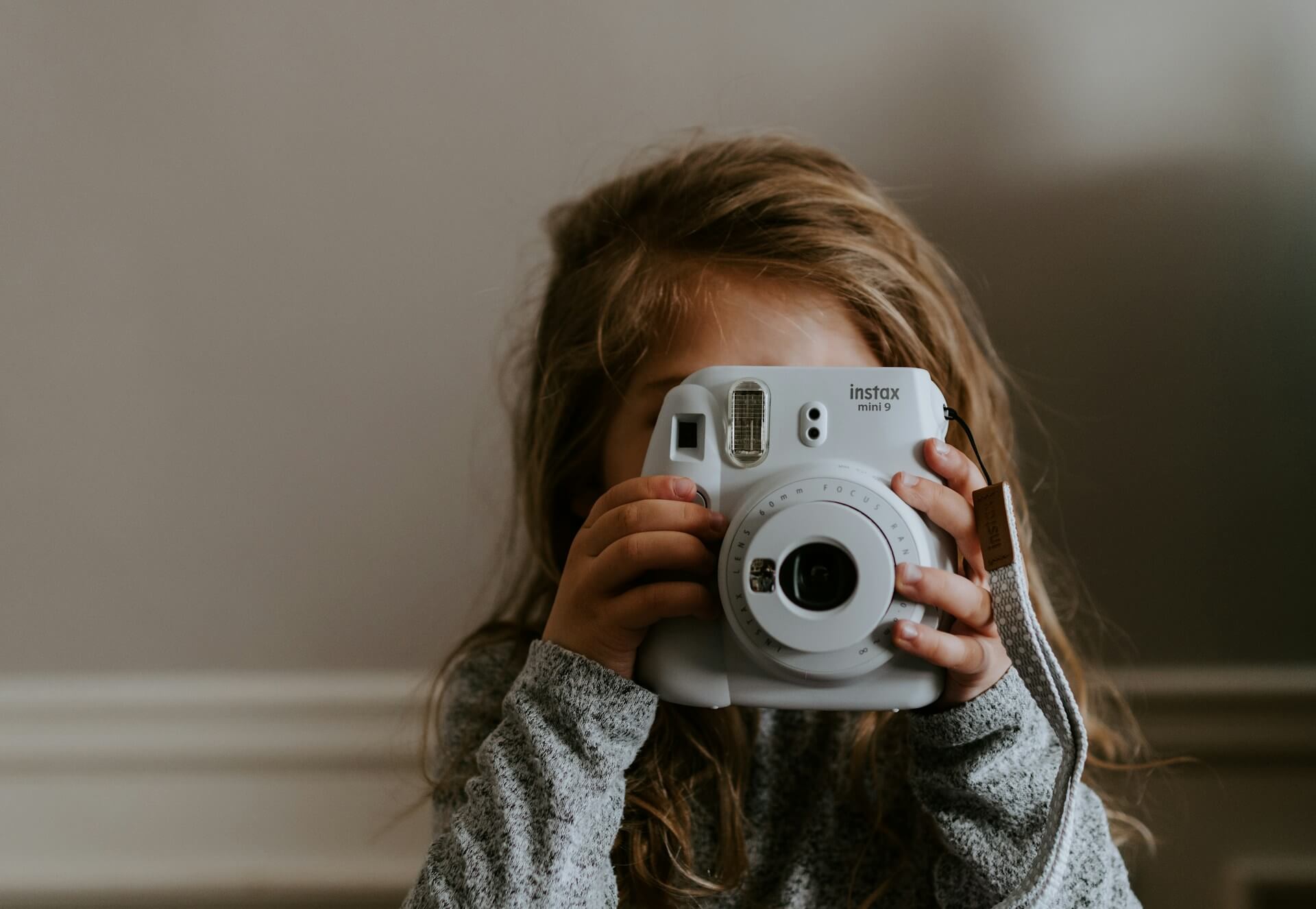 Kislány egy jégfehér Instax Mini 9 fényképezőgéppel. Hosszú hajú kislány, aki egy jégfehér Fujifilm Instax Mini 9 instant fényképezőgéppel takarja el az arcát, mintha fotózna.