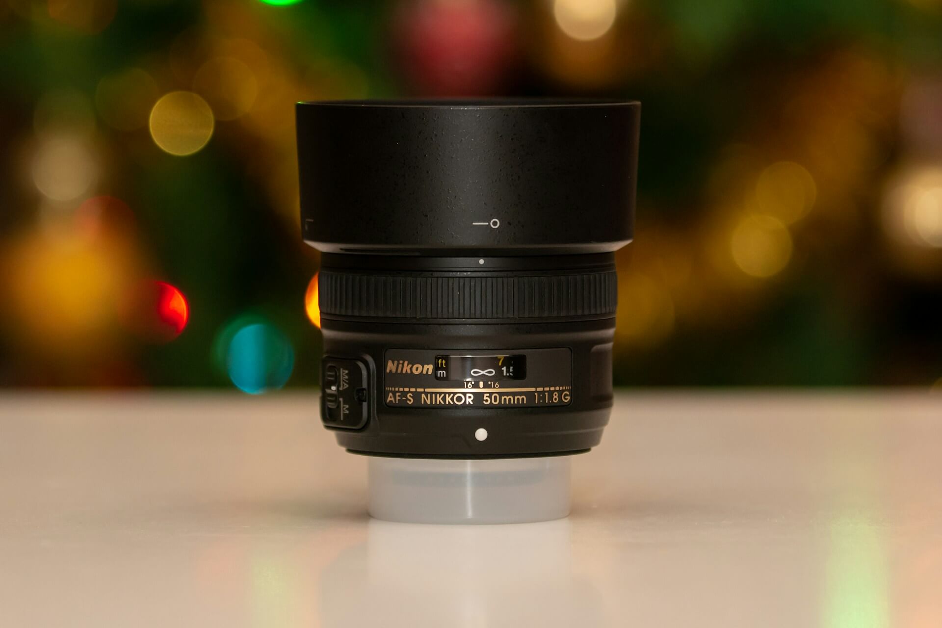 Egy fekete Nikon AF-S NIKKOR 50mm f/1.8G objektív napellenzővel egy világos felületen, háttérben ünnepi fények lágy elmosódásával (bokeh).