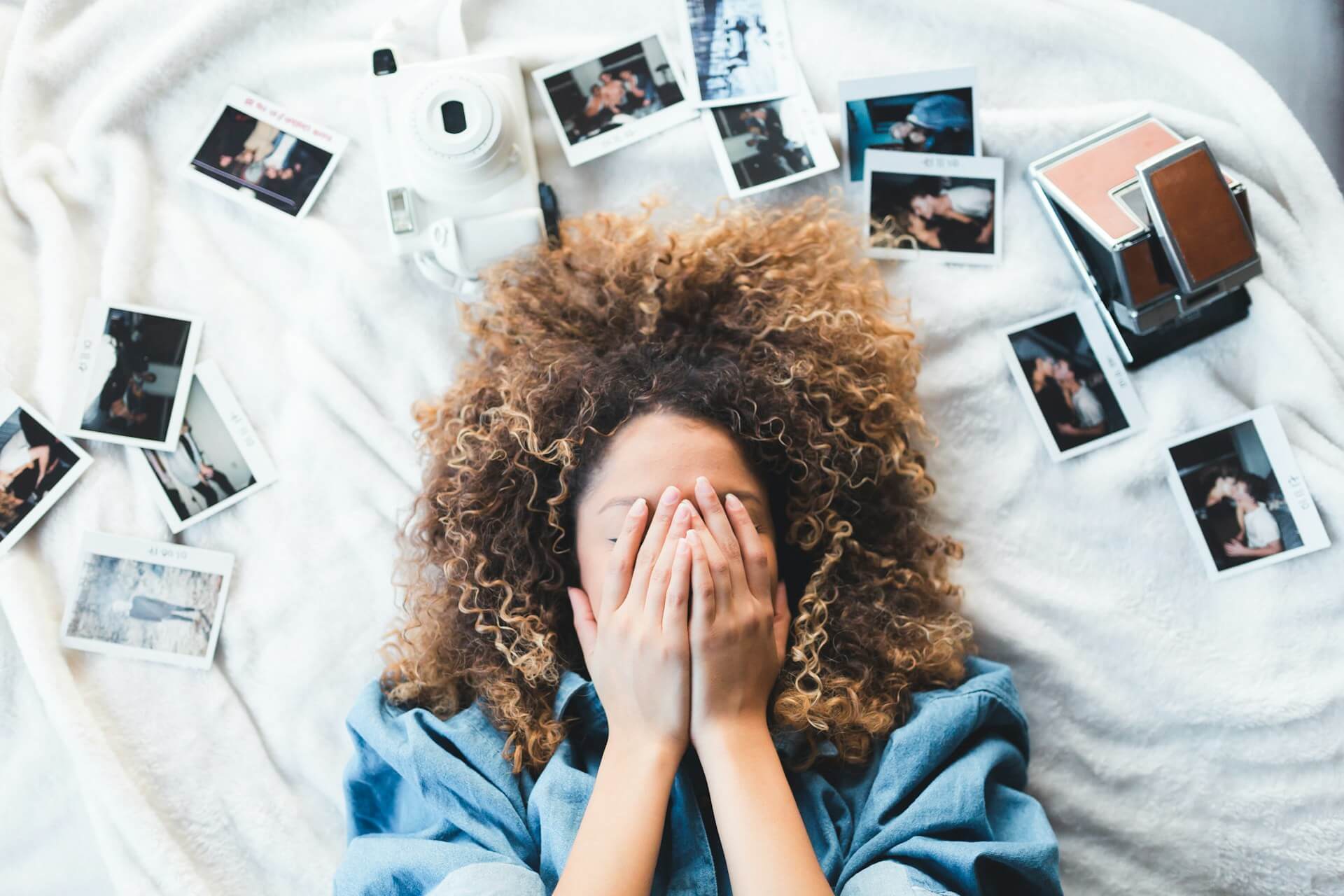 elülnézeti kép egy göndör hajú nőről, aki az arcát eltakarva fekszik egy fehér takarón, szétszort Instax fotók és fényképezőgépek között.