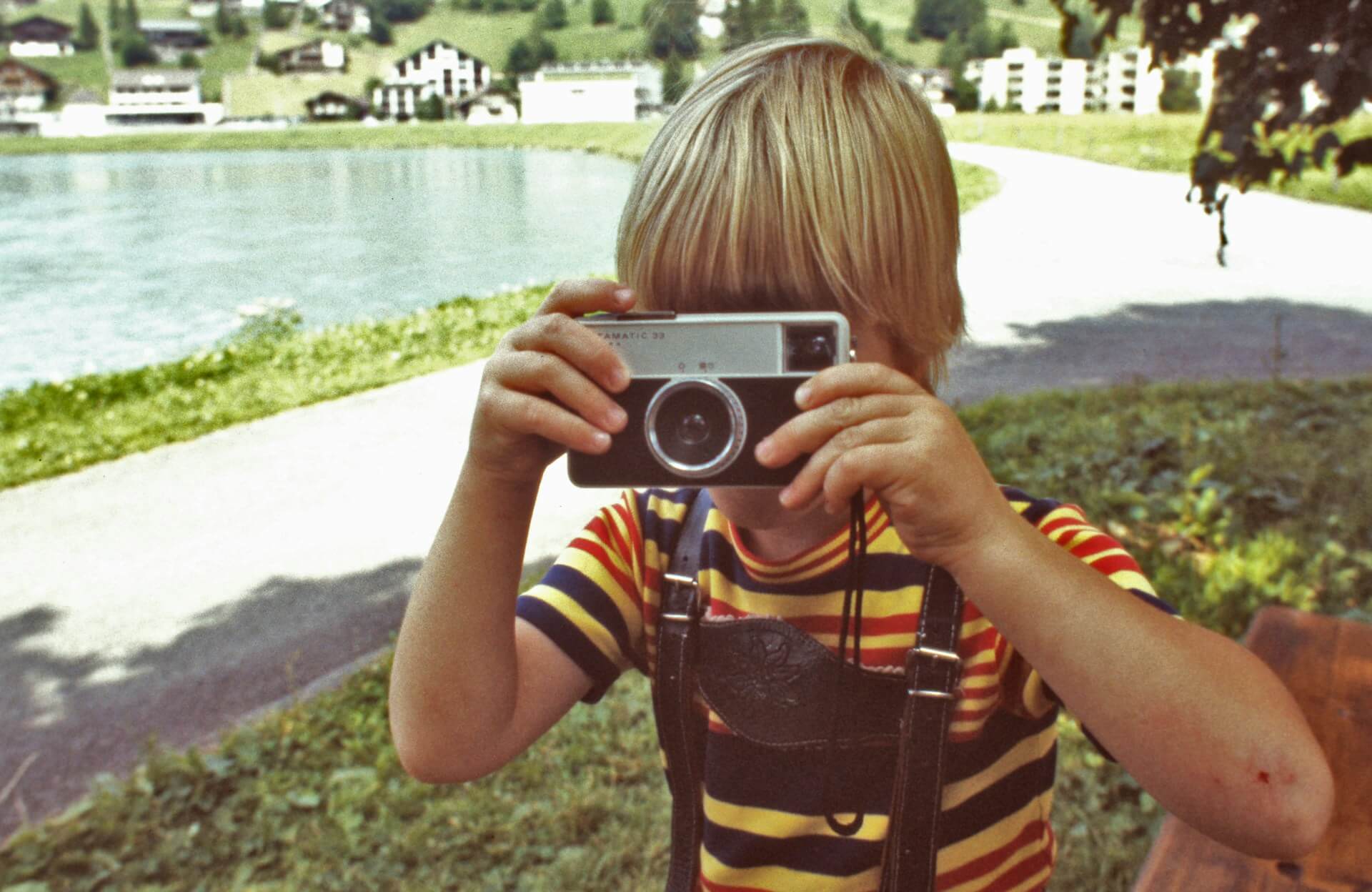 Gyermek egy vintage fényképezőgéppel a tengerparti vagy tóparti természetben. Csíkos pólós, szőke kisfiú egy retro analóg fényképezőgépet tart a szeme elé egy tóparti, hegyvidéki tájban.