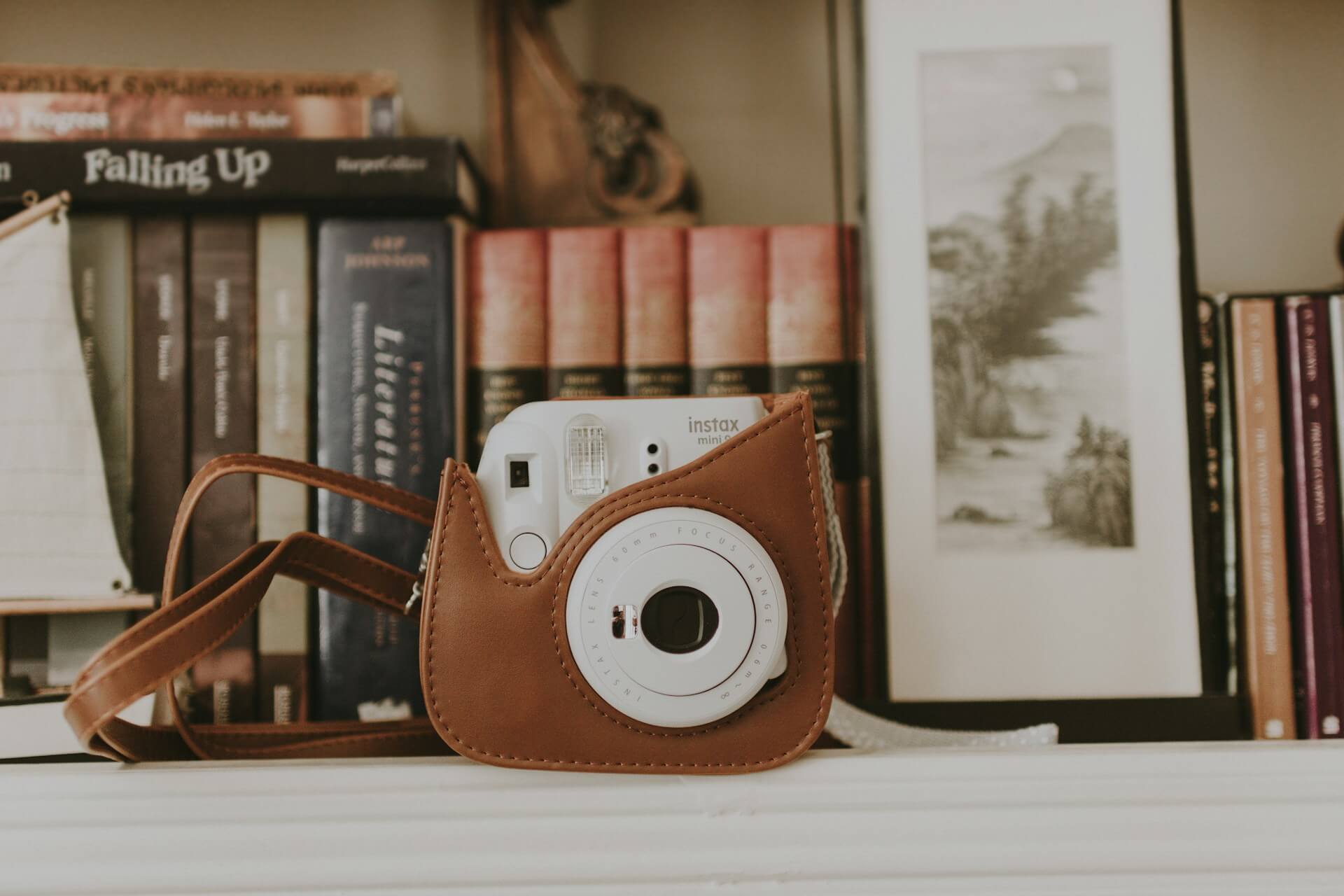 Fehér Fujifilm Instax Mini fényképezőgép barna bőrtokban, könyvespolcon elhelyezve, könyvek és egy keretezett kép előtt.