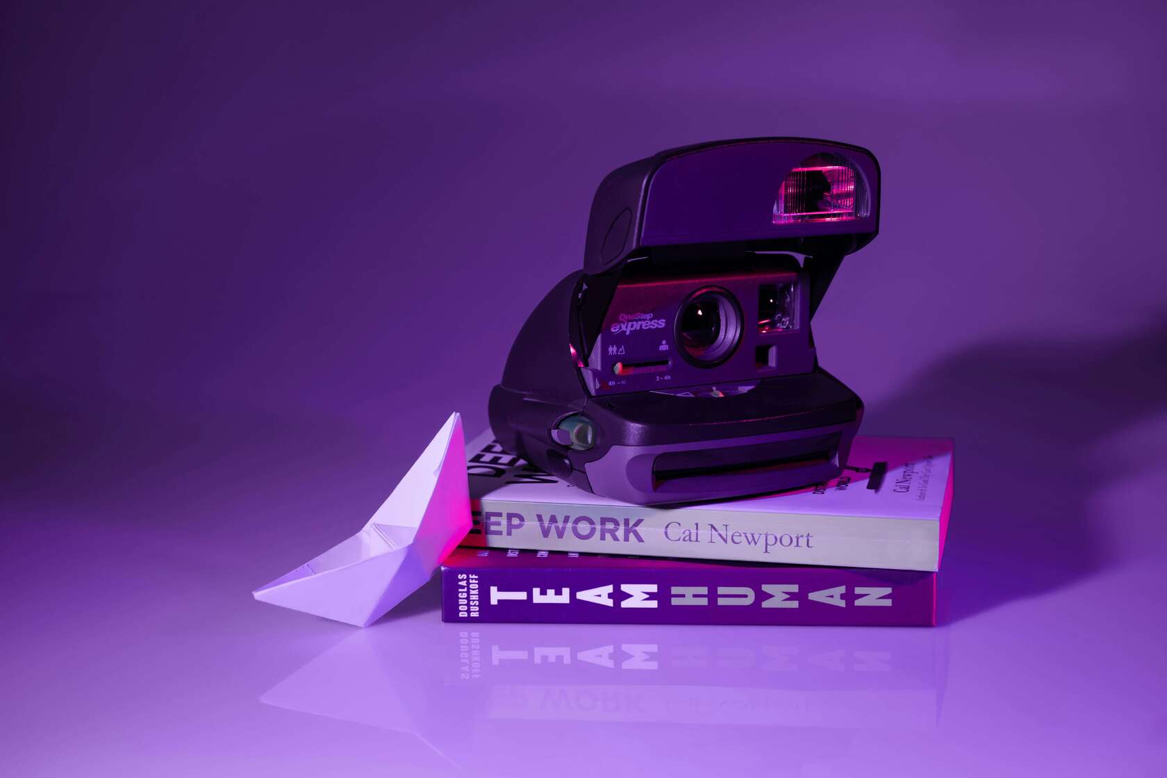 Polaroid fényképezőgép neonfényben Lila és rózsaszín fényekkel megvilágított Polaroid fényképezőgép könyvekre helyezve, mellette papírhajóval.