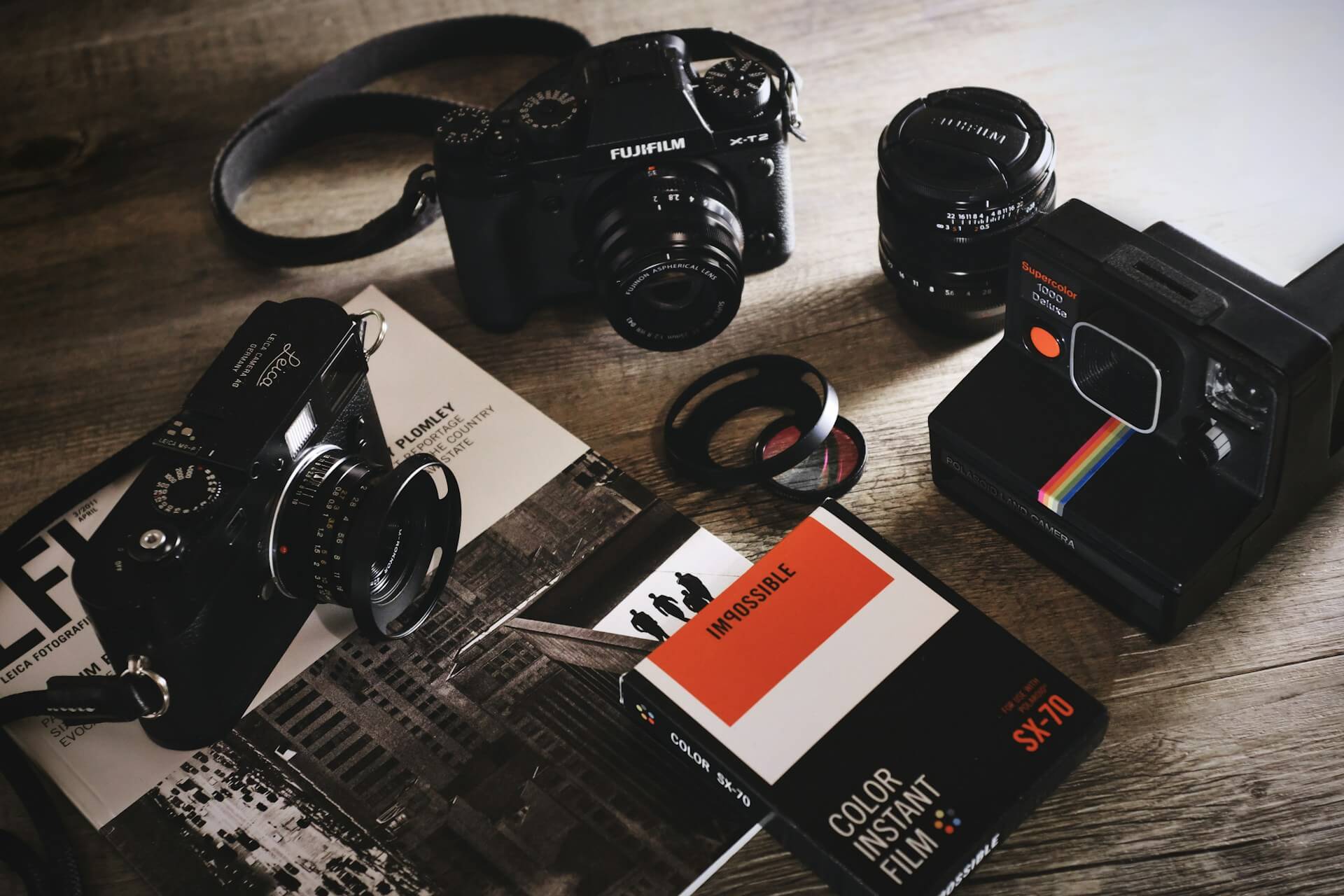 Analóg és digitális fényképezőgépek, objektívek és instant film egy faíróasztalon. Felülnézeti fotó egy fajellegű asztalról, amin Leica és Fujifilm vázak, objektívek, Polaroid fényképezőgép és Impossible SX-70 instant film látható.