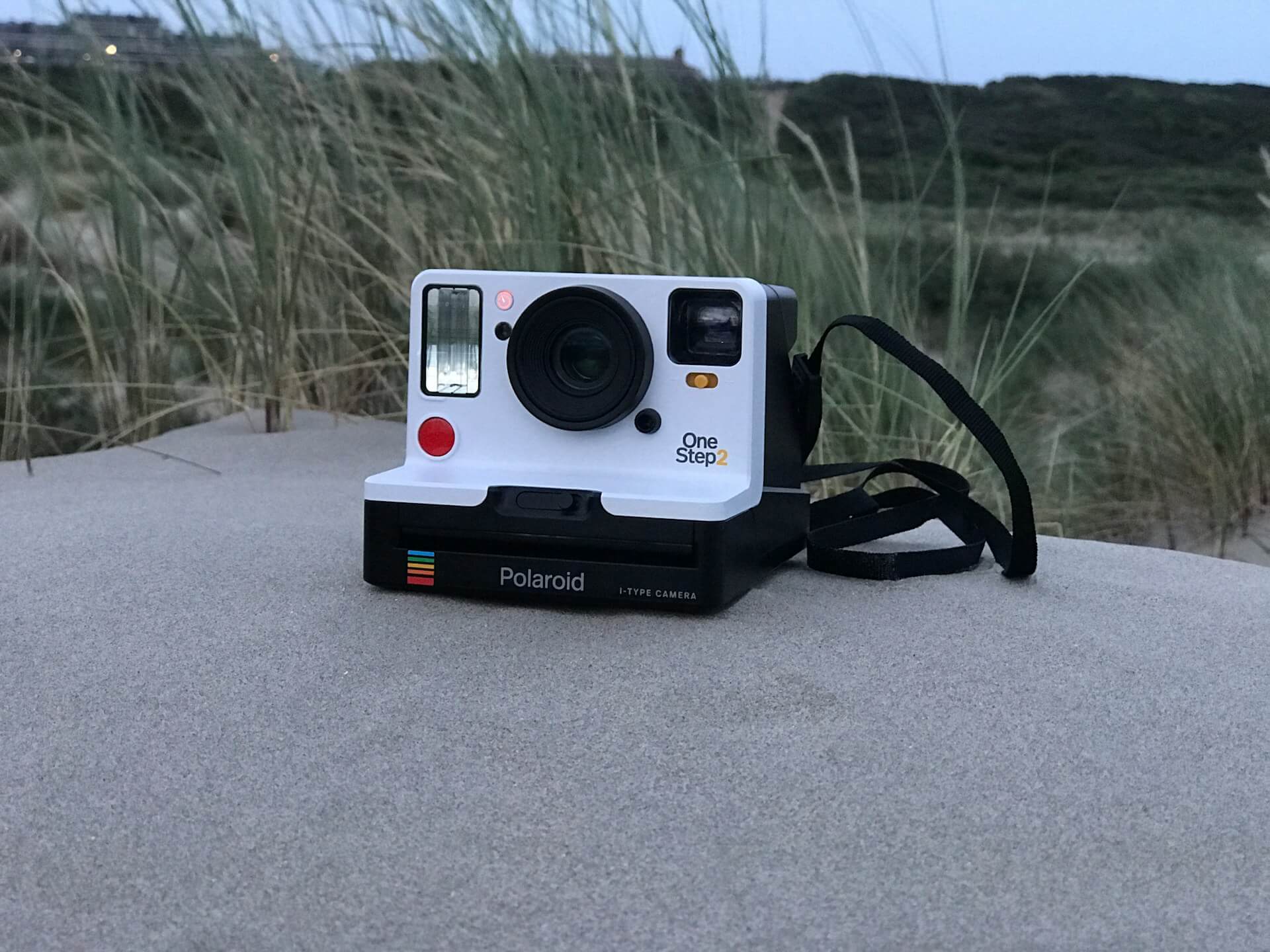 Fehér és fekete Polaroid OneStep 2 i-Type instant fényképezőgép a homokban, háttérben tengerparti fűvel.