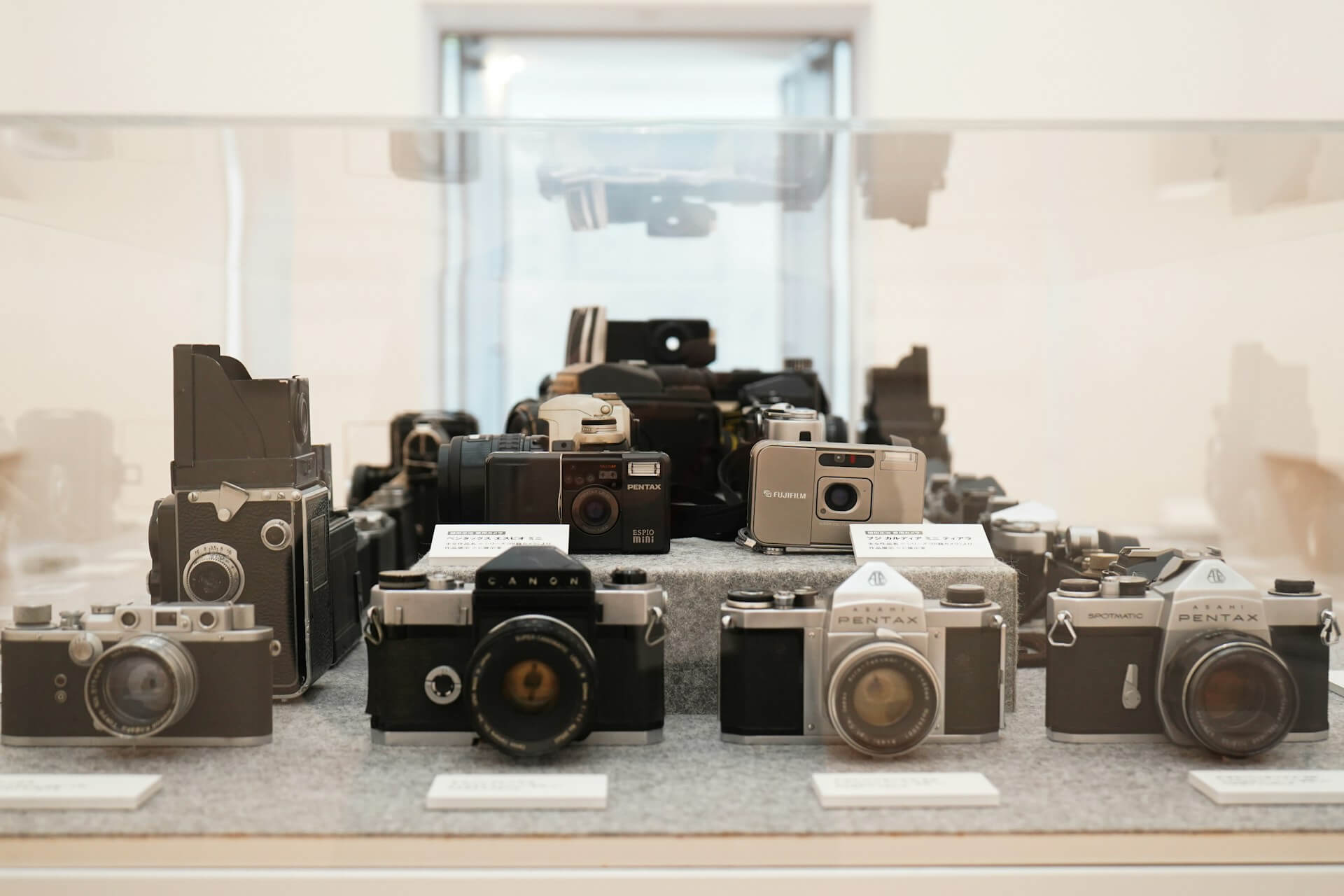 Különböző vintage Pentax, Canon és Fujifilm fényképezőgépek kiállítva egy múzeumi üvegvitrinben, japán nyelvű információs kártyákkal.